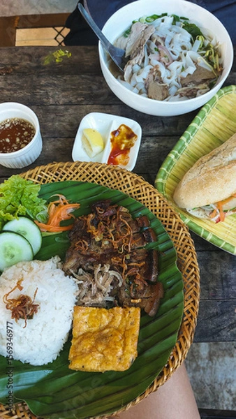 Fototapeta Authentic traditional local Vietnamese dishes featuring trung chien or egg omelet, thit heo nuong xa or grilled pork chop and rice on a banana leaf alongside pho bo or beef noodle soup and baguette