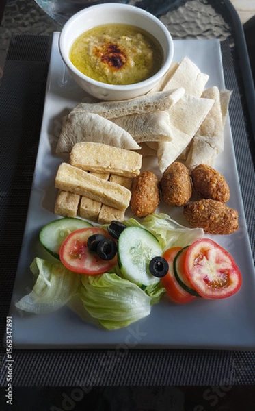 Fototapeta Mediterranean vegan appetizer platter with hummus, pita, falafel, fried tofu and fresh vegetables on a white plate, healthy diet and wellness