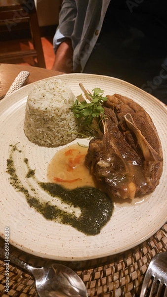 Fototapeta A plate with lamb chop, herbed rice, and pesto on a wooden table with a woven mat, close up overhead view