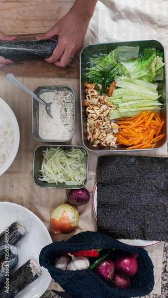 Fototapeta A vertical arrangement of ingredients while hands are rolling a gimbap or seaweed rice roll near vegetables and a sauce bowl