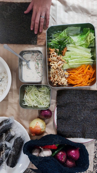 Fototapeta Hands set to prepare use seaweed sheet, surrounded by colorful vegetables, rice and other ingredients for making kimbap, traditional south korean food