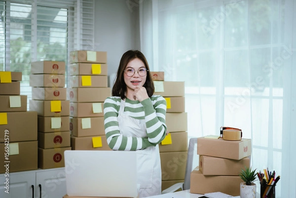 Obraz A young woman entrepreneur manages her online business from home, surrounded by parcel boxes, using a laptop, calculator, and notepad to handle shipping and inventory.