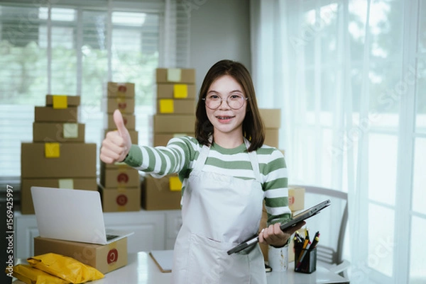 Obraz A young woman entrepreneur manages her online business from home, surrounded by parcel boxes, using a laptop, calculator, and notepad to handle shipping and inventory.
