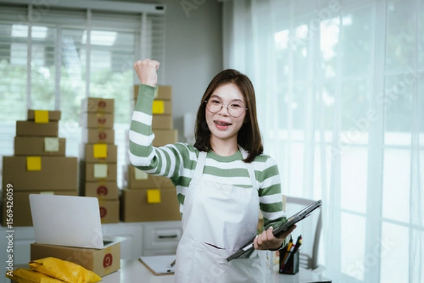 Obraz A young woman entrepreneur manages her online business from home, surrounded by parcel boxes, using a laptop, calculator, and notepad to handle shipping and inventory.