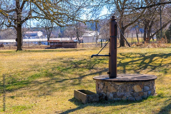 Fototapeta Rustic water pump in countryside with stone base and trees