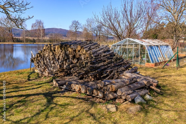 Fototapeta Woodpile beside lake with greenhouse and scenic landscape