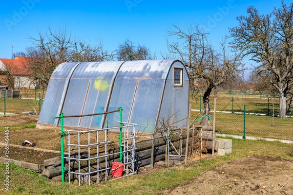 Fototapeta Sunny greenhouse in rural garden setting with bare trees and blue sky