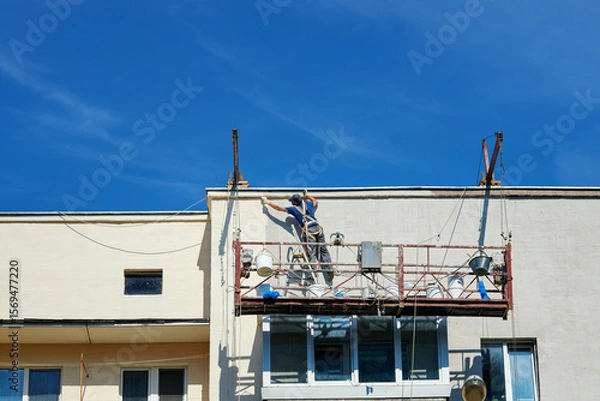 Fototapeta Unrecognizable construction worker on suspended scaffolding renovating facade of an apartment building, exterior maintenance for urban infrastructure improvement. Man restoring and painting facade