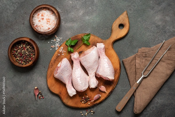 Fototapeta Raw chicken drumsticks on round wooden cutting board with spices flat lay on dark grunge background, top view