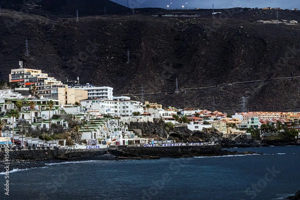 Obraz Houses and buildings in the town of Panta del Hidalgo with an inscription on concrete barriers on the shore of the Atlantic Ocean on the island of Tenerife with a mountain in the background	

