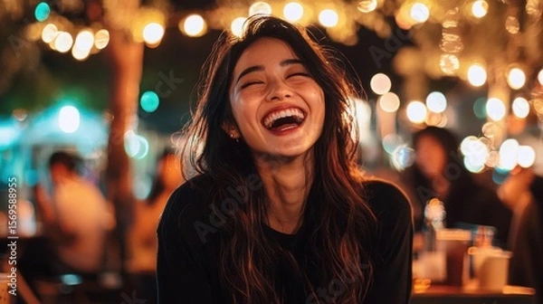 Obraz Close-up portrait of a young woman with long dark hair, laughing with her eyes closed and her mouth wide open. she is wearing a black top and appears to be in a restaurant or bar.