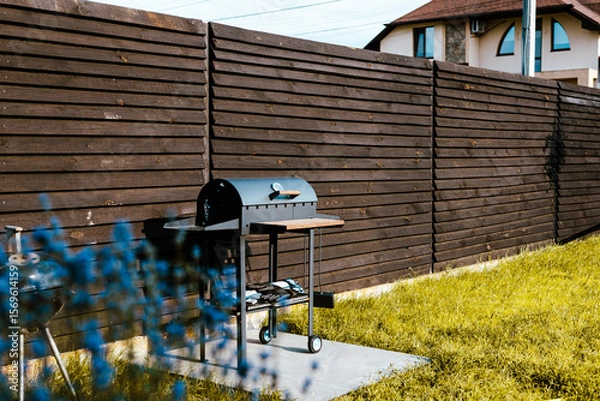 Fototapeta A barbecue grill and patio are ready for outdoor cooking in a backyard. A BBQ grill in front of a wooden fence in a backyard. The backyard also has grass and a house in the background. Leisure weekend