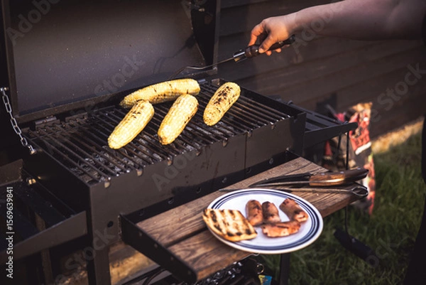 Fototapeta Grilled corn on the cob and sausages are ready for a summer barbecue party. A person grilling corn on the cob and sausages on a barbecue grill outdoors on a wooden table, enjoying a summer meal.