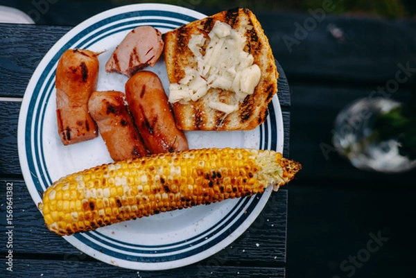 Fototapeta Grilled sausages, corn, and bread arranged on a white plate. A delicious plate of grilled food, perfect for a summer barbecue. Includes sausages, corn on the cob, and bread.