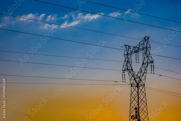 Fototapeta High voltage power towers and lines under the background of blue sky, white clouds, and orange sunlight