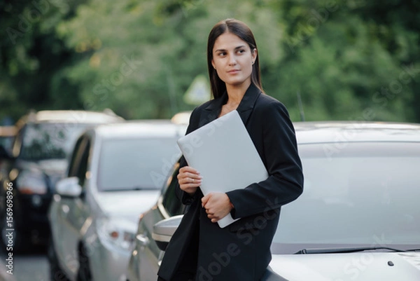 Obraz Woman in business suit holding laptop leaning on her car