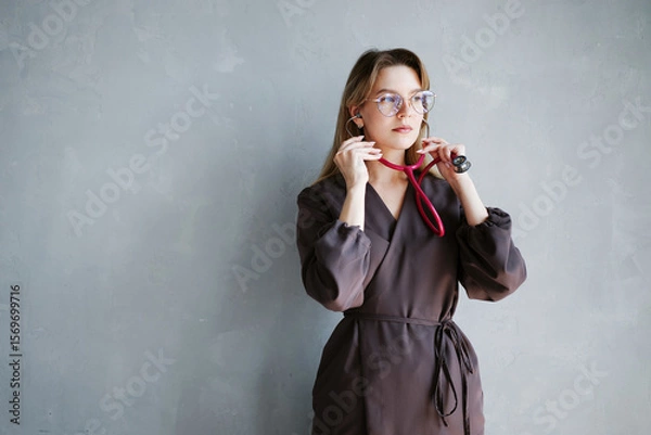 Fototapeta Portrait of female doctor in dark medical uniform putting on stethoscope while standing against gray wall.