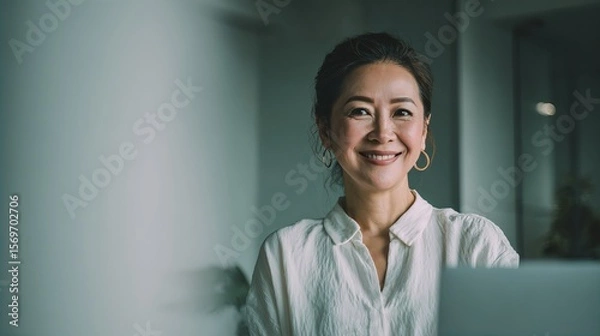 Obraz A smiling woman with dark hair pulled back, wearing a white blouse and gold hoop earrings, sits in a softly lit indoor space, exuding warmth and confidence.