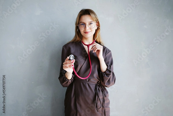 Fototapeta Portrait of female doctor in dark medical uniform using stethoscope and smiling at camera against gray background.