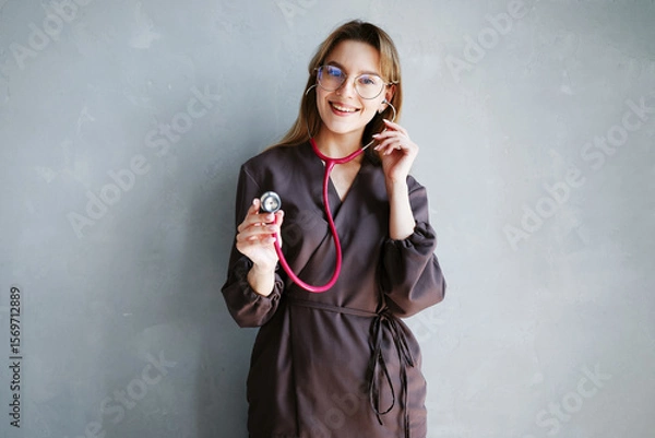 Fototapeta Portrait of female doctor in dark medical uniform smiling and using stethoscope while standing against gray wall.