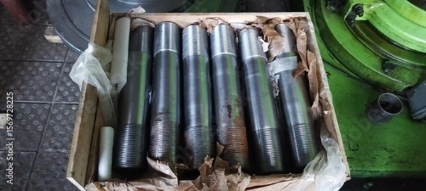 Fototapeta A crate of newly manufactured flange studs, steel threaded rods, with industrial machinery in the background. Close-up of metal flange studs, packaged in a wooden box for transport. Metal studs 