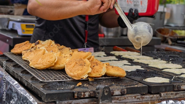 Obraz Taiyaki being prepared at street food stall with traditional griddle. Outdoor culinary scene with cooks in action.