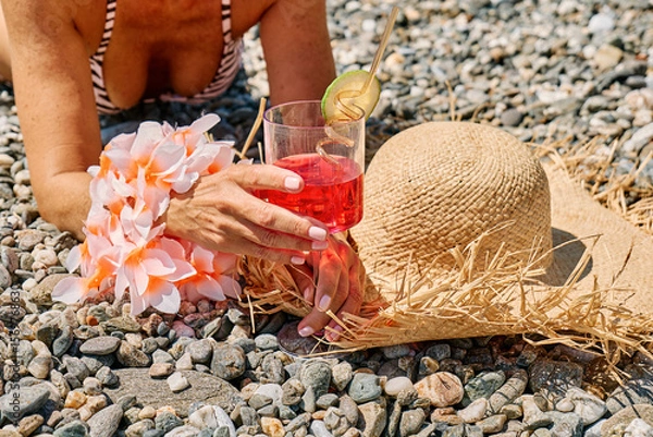 Fototapeta Anonymous woman in bikini ad straw hat holding summer refreshing red soda drink on pebble beach.