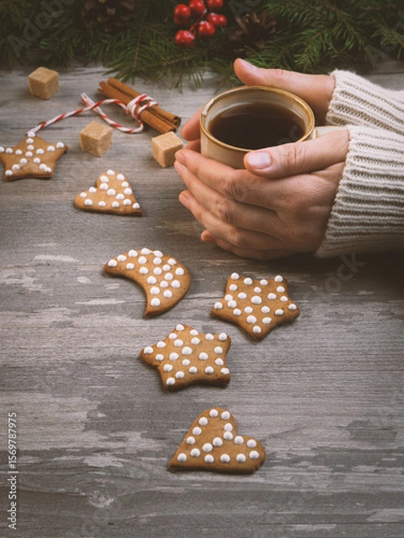 Fototapeta Christmas atmospheric set. Female hands in a sweater with coffee mug among tradition gingerbreads and holidays decoration on a wooden background, vertical image.