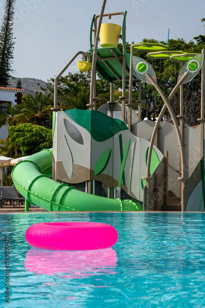 Fototapeta A pool with a slide and a pink rubber ring floating in the water. The pool is surrounded by trees and has a jungle theme