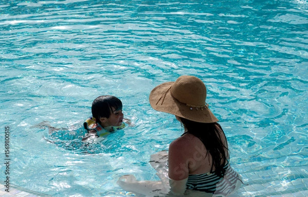 Fototapeta A woman is sitting in a pool with a child. The woman is wearing a straw hat and the child is wearing a yellow life jacket. Scene is lighthearted and fun, as the woman