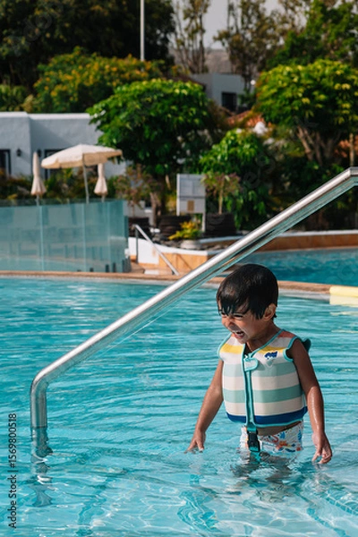 Fototapeta A young boy is in the water wearing a life jacket. He is standing in the pool and looking up at the camera