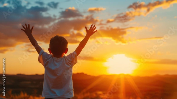 Fototapeta A young boy is standing in a field with his hands raised in the air