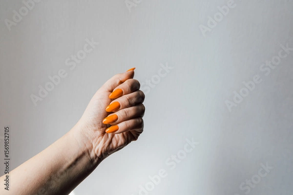 Obraz Hand with bright orange nails holds a fist against a plain white background