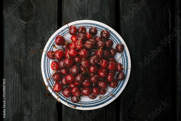Fototapeta A vibrant image of ripe cherries on a plate set against a dark wooden backdrop. A plate of fresh, juicy cherries sits on a rustic black wooden table, capturing a vibrant, appetizing scene.