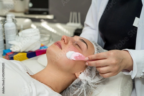 Fototapeta Beautician applies cleansing and moisturizing white cream with help of brush on client's face
