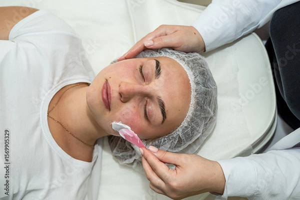 Fototapeta Beautician applies cleansing and moisturizing white cream with help of brush on client's face