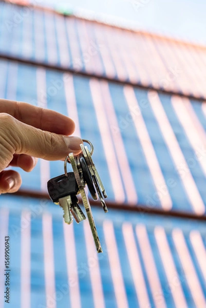 Fototapeta Hand Holding Keys in Front of Modern Glass Office Building. A close-up image of a hand holding a set of metallic keys against the background of a sleek glass skyscraper