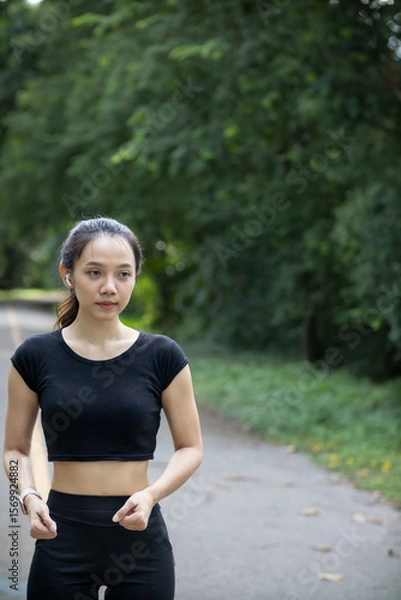 Fototapeta Portrait of a beautiful Asian woman in a black sportswear walking alone on the street after jogging in the morning. The athlete in a black is walking to warm up her body before jogging to get healthy.