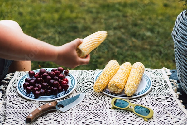 Fototapeta A summer picnic scene featuring corn on the cob, cherries, and vintage green sunglasses. Close-up of a person enjoying a summer lunch or brunch outdoors with corn on the cob, a knife. Summertime fun.