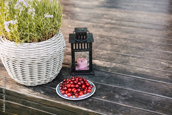 Fototapeta A delightful still life showcasing cherries, lavender, and a candle lantern on a wooden terrace. A rustic scene features a basket of lavender, red berries on a weathered wooden surface on a backyard.