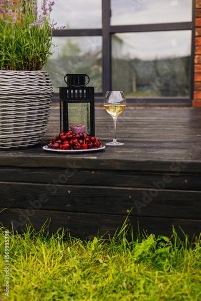 Fototapeta Enjoying a glass of white wine with fresh red cherries in a relaxing outdoor setting. A rustic outdoor setting showcases a glass of white wine, cherries, and a lantern on a wooden deck.