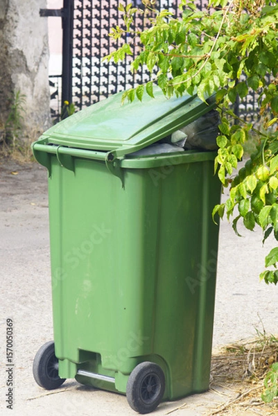 Fototapeta Full green plastic garbage bin on wheels on sidewalk ready for removal by municipal service. Urban waste overflow and outdoor trash disposal. Environmental problem in city with litter and pollution