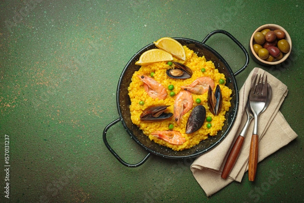 Obraz Seafood paella with rice, shrimp, mussels, peas, and lemon wedges served in a black traditional pan on a green rustic background with olives and cutlery, copy space