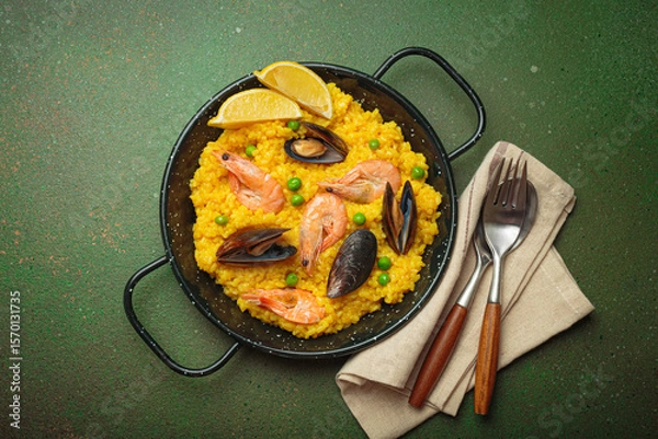 Obraz Seafood paella with rice, shrimp, mussels, peas, and lemon wedges served in a black traditional pan on a green rustic background with olives and cutlery