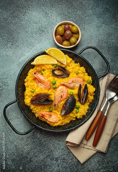 Obraz Seafood paella with rice, shrimp, mussels, peas, and lemon wedges served in a black traditional pan on a dark rustic background with olives and cutlery