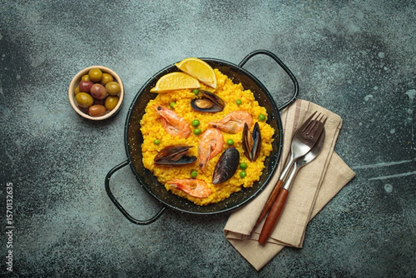 Obraz Seafood paella with rice, shrimp, mussels, peas, and lemon wedges served in a black traditional pan on a dark rustic background with olives and cutlery