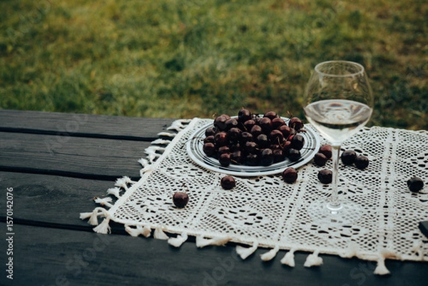 Fototapeta A glass of white wine and a plate of cherries on a table outside on a sunny day. A refreshing glass of white wine alongside a plate of ripe cherries on a table outdoors, ready for enjoyment.