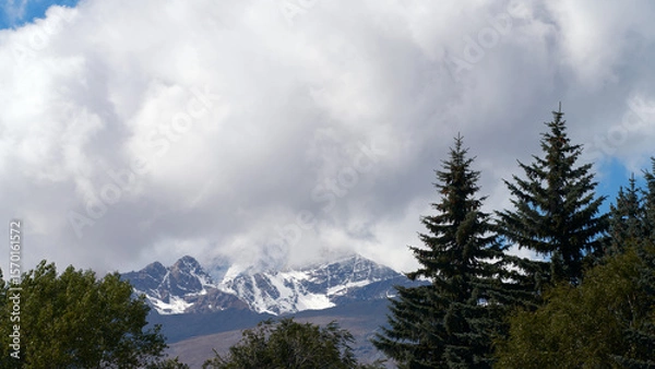 Fototapeta High mountain landscape. Snow-capped peak of rocky mountain in cloud against background of mountain spruce and other trees.