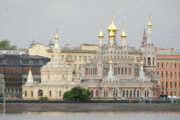 Fototapeta Skorbyashchenskaya Church in Saint Petersburg, featuring ornate architecture and golden onion domes, located by the riverside against historic buildings.