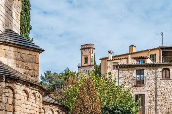 Fototapeta Medieval stone architecture of Girona with decorative tower, Catalan buildings, terracotta roofs, and arched details nestled in greenery under a clear blue sky, capturing the Spain historic charm